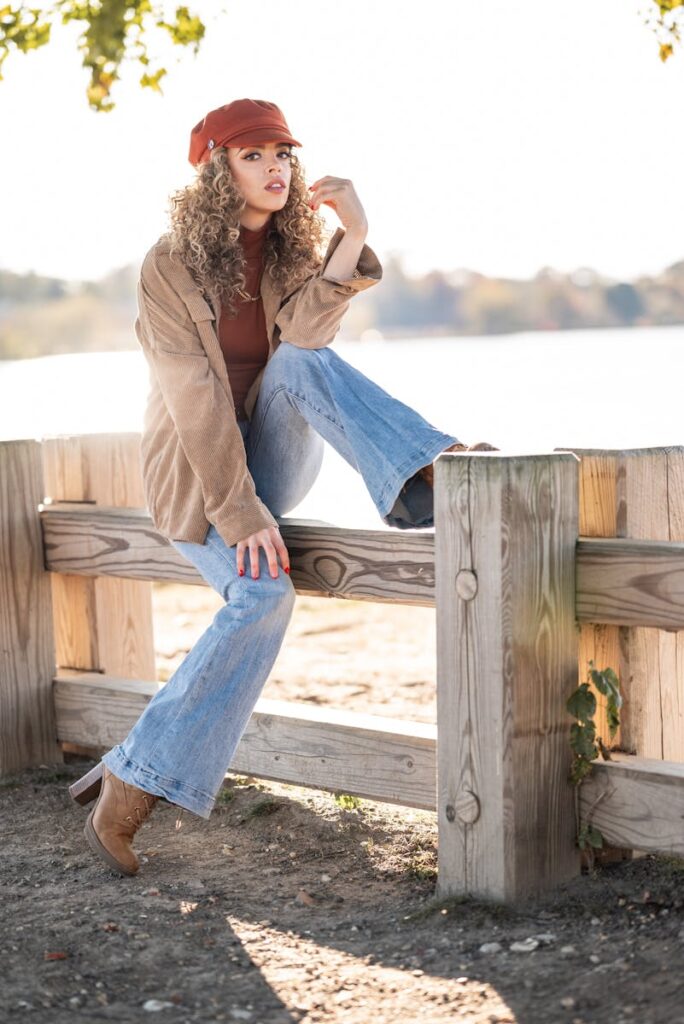 A fashionable woman poses by a lake in autumn, wearing jeans, jacket, and hat.