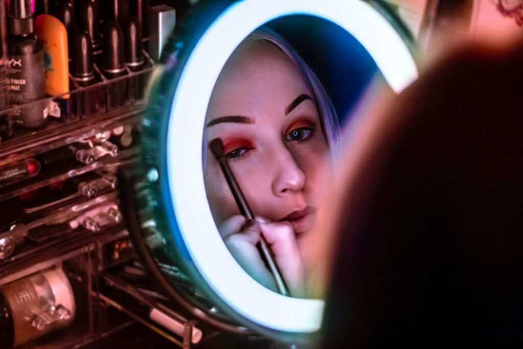 A woman carefully applies eye makeup while looking into a ring-lit mirror in a studio setting.