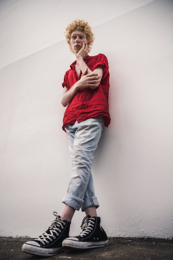 Stylish young man with curly hair posing outdoors against a white wall in a casual outfit.