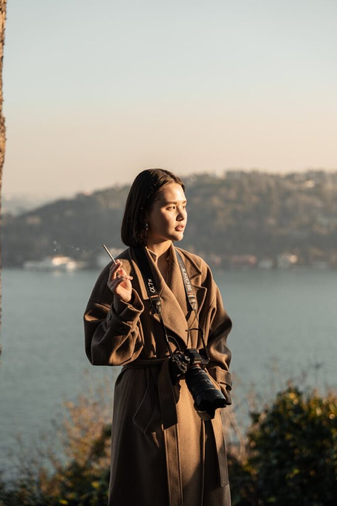 Woman in brown trench coat with camera and cigarette by a serene lakeside view.