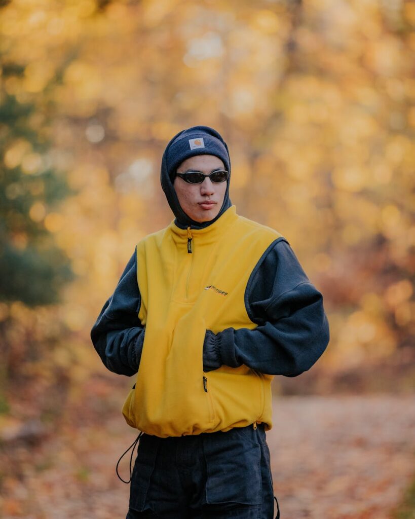 Man wearing a yellow jacket and beanie, posing outdoors in an autumn setting.
