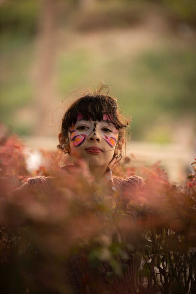 A young girl with colorful face paint stands amidst foliage outdoors, radiating joy.