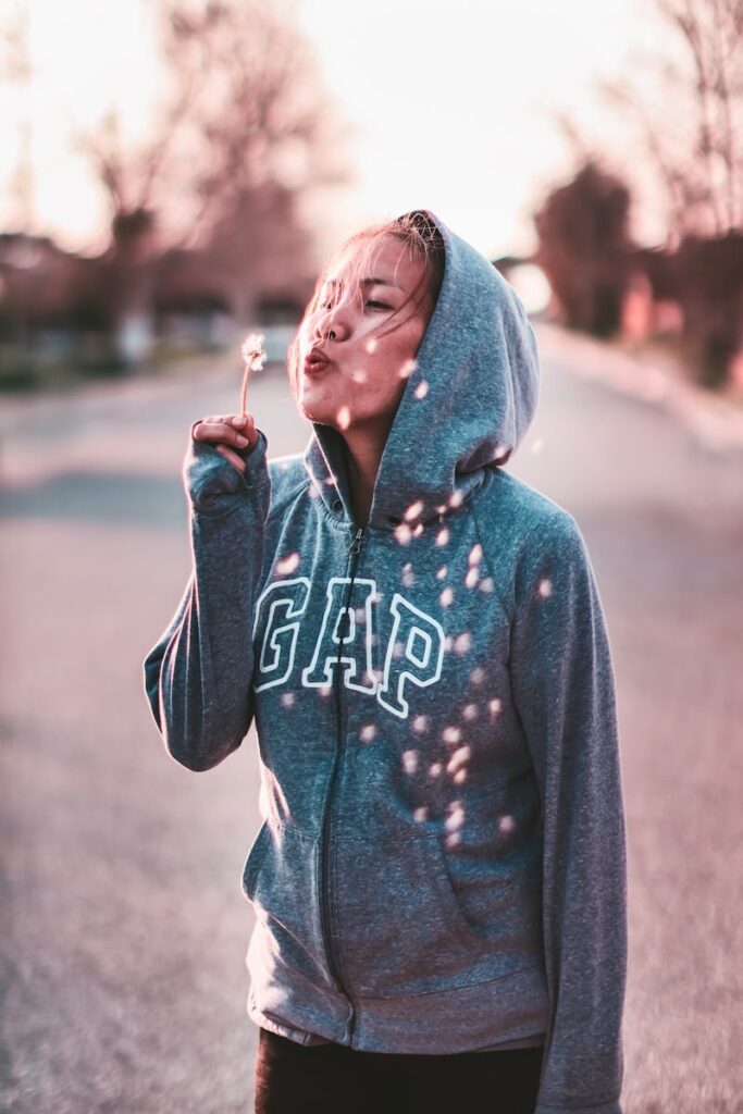 A young woman wearing a hoodie blowing upon a dandelion outdoors, creating a whimsical scene.
