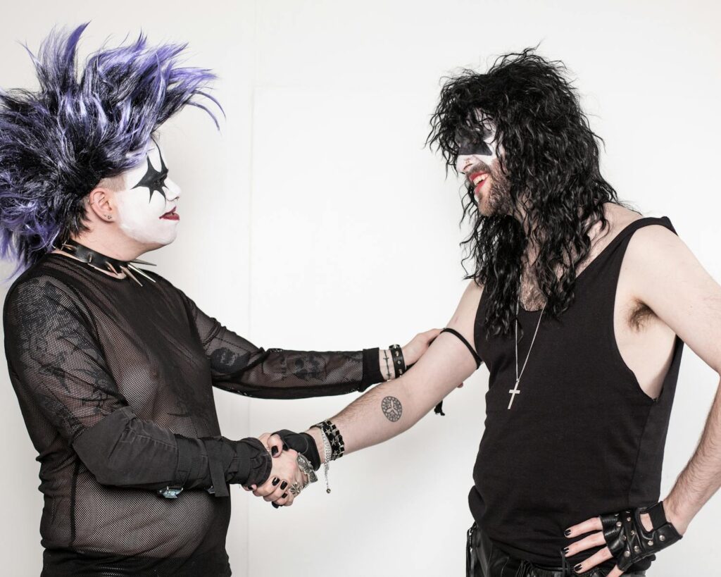 Two friends in goth attire and makeup shaking hands with a white background.