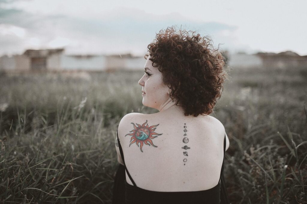 Curly-haired woman with back tattoo sitting in a grassy field.