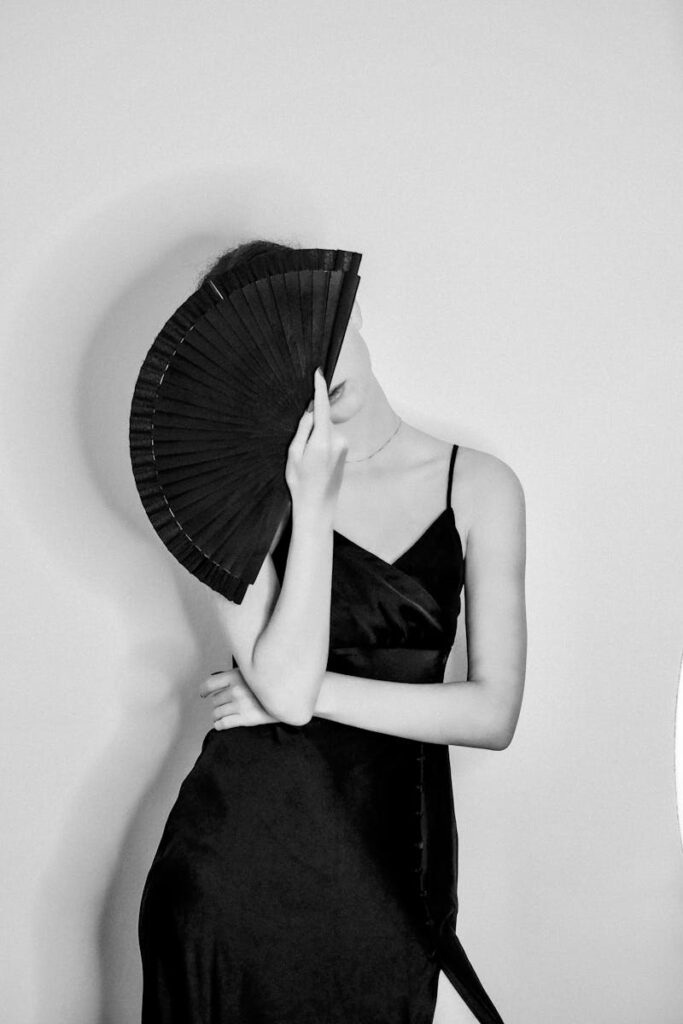 Black and white photo of a woman in a dress posing with a fan, creating a chic and mysterious atmosphere.