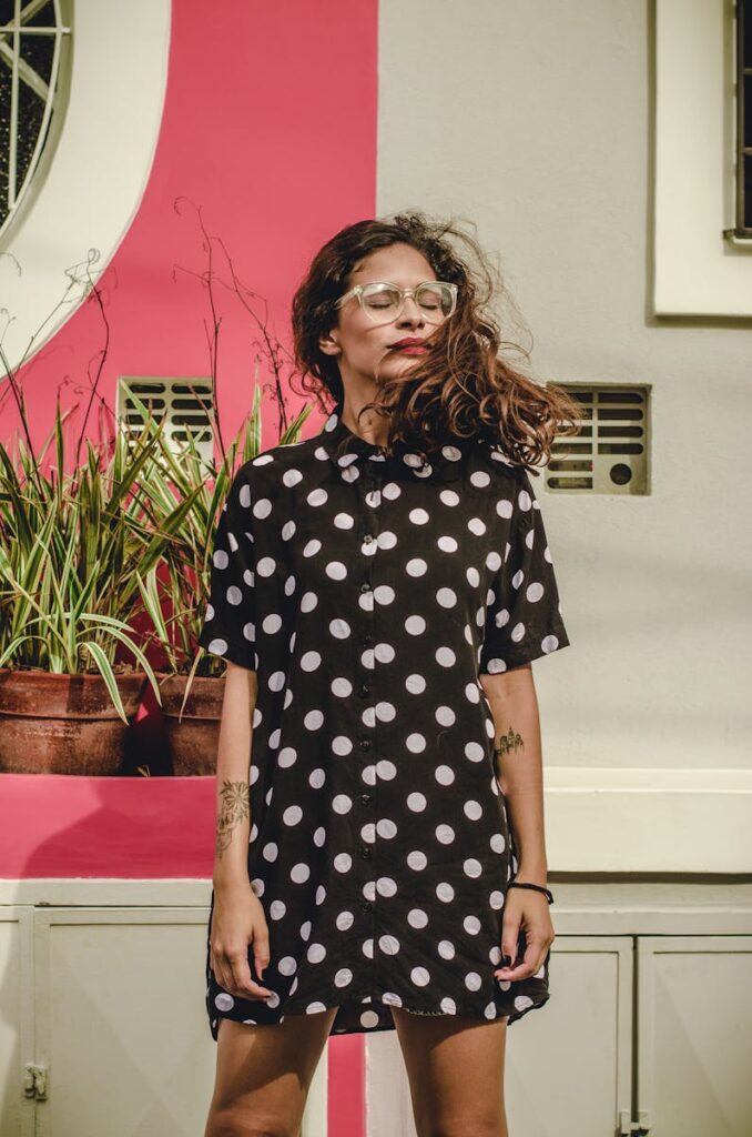 Trendy woman in a polka dot dress posing confidently with a pink background.