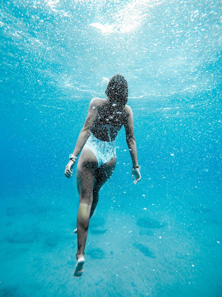 Woman swimming underwater with bubbles in blue ocean, captured from behind.