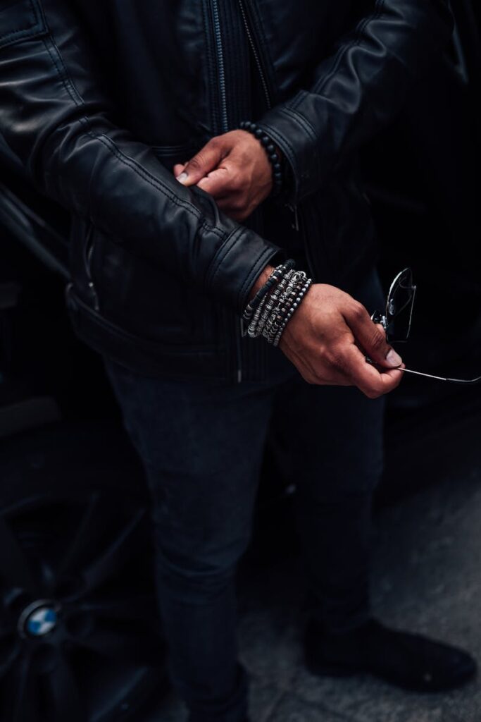 Man in a black leather jacket wearing bracelets, holding sunglasses, showcasing fashion style.