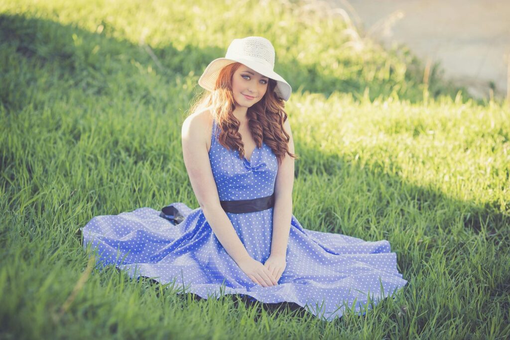 A young woman in a polka dot dress and sun hat sits on a grassy field, enjoying a sunny day.