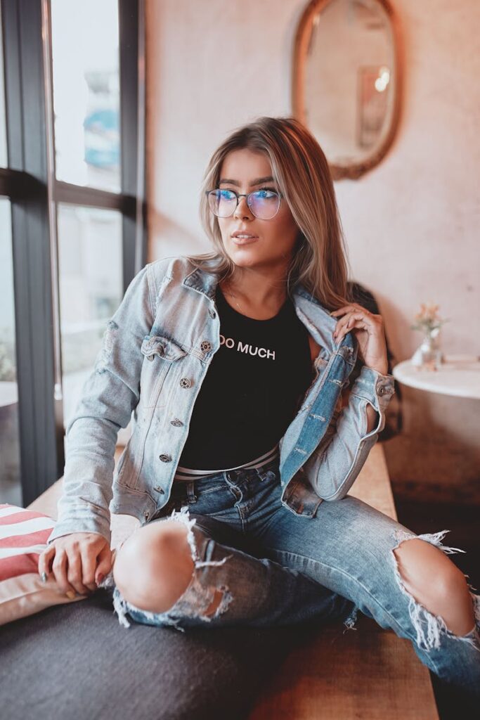 Trendy woman sitting indoors wearing denim jacket and glasses, emanating modern fashion vibes.
