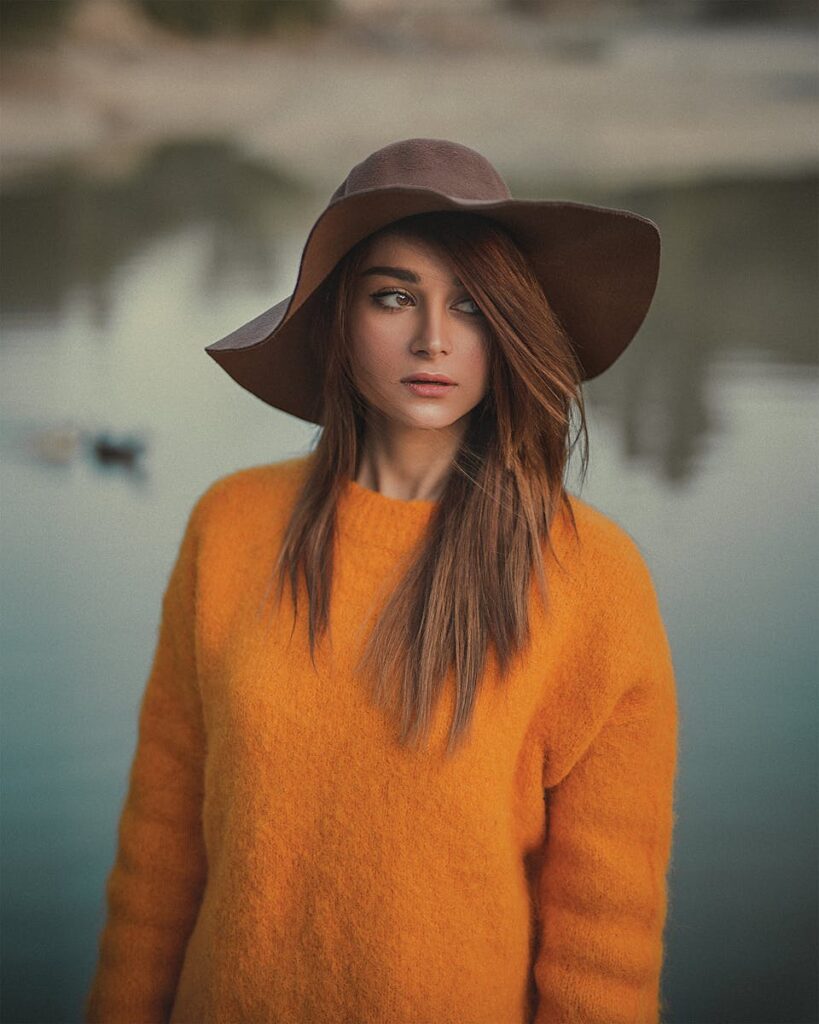 Woman in an orange sweater poses by a serene body of water in Paris.