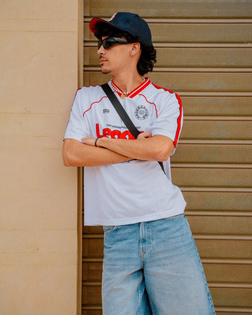 Stylish man wearing a cap, sunglasses, and casual jeans stands confidently outdoors.