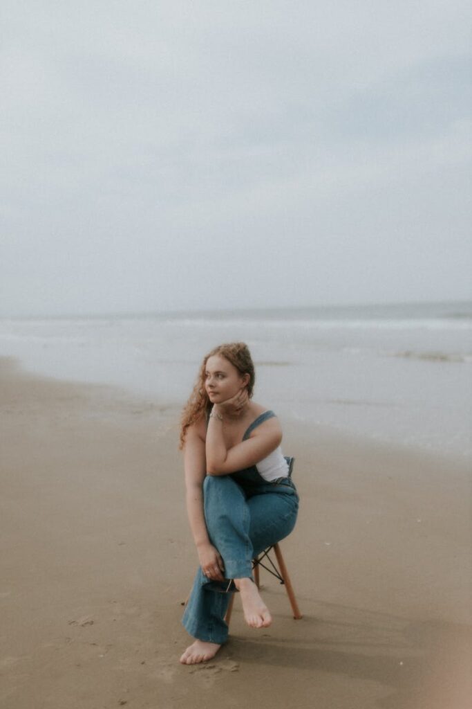 A woman in denim overalls sits on a chair at the beach, exuding a calm and relaxed vibe.