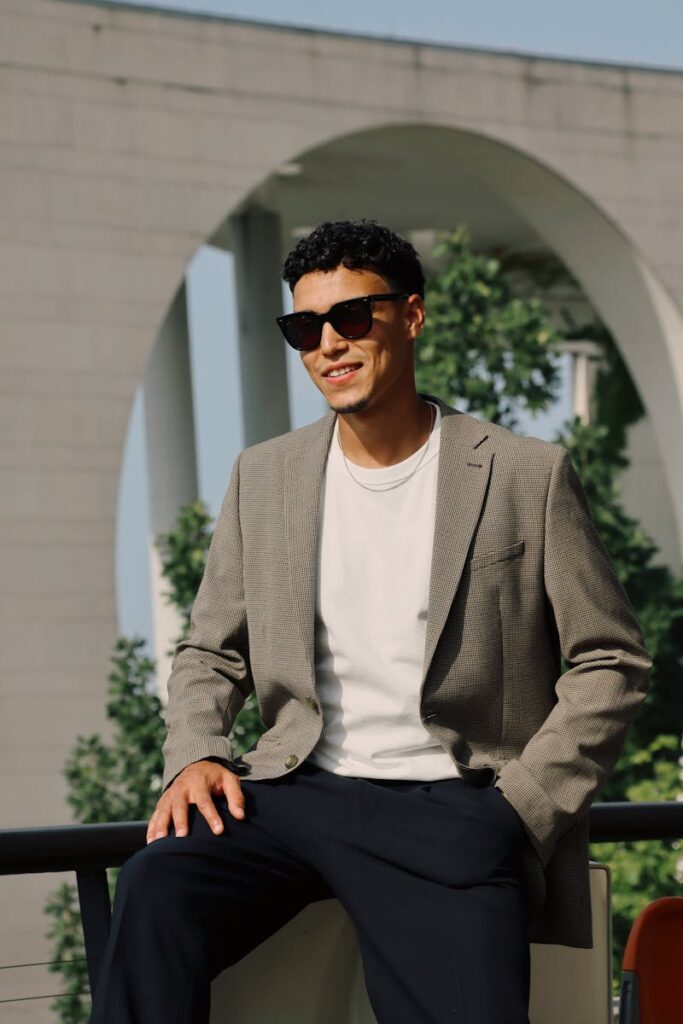 Fashionable man in sunglasses and blazer sitting outdoors in Berlin. Urban elegance captured.