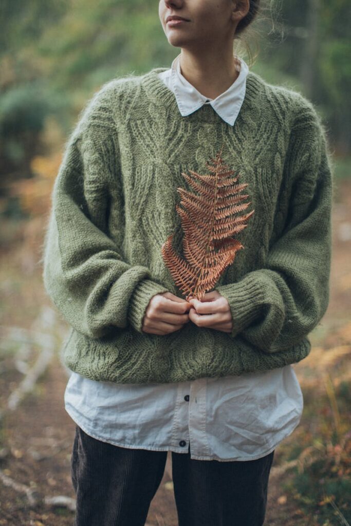 A young woman outdoors in Scotland, wearing a green knit sweater and holding an autumn leaf, exuding warmth and style.