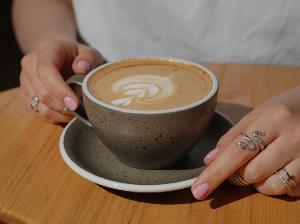 A close-up shot of hands with rings holding a cappuccino featuring delicate latte art on a wooden table.