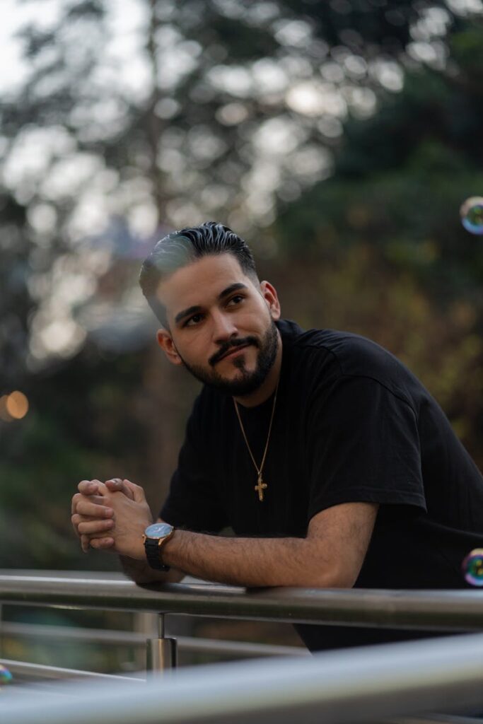 Portrait of a thoughtful man with a beard and gold cross pendant leaning on a balcony railing.