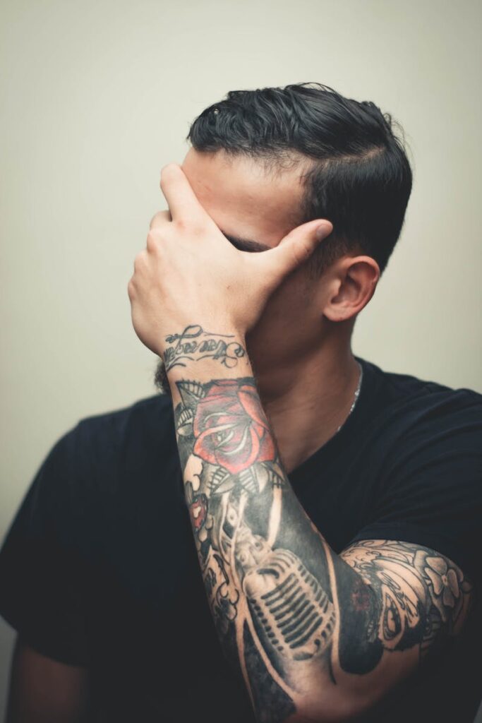 Stylish man with detailed arm tattoos covers face with hand, showcasing intricate body art.