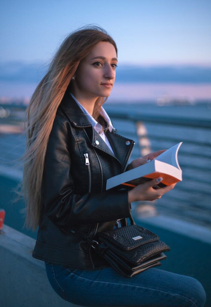 Woman in black leather jacket reading by the water during sunset.