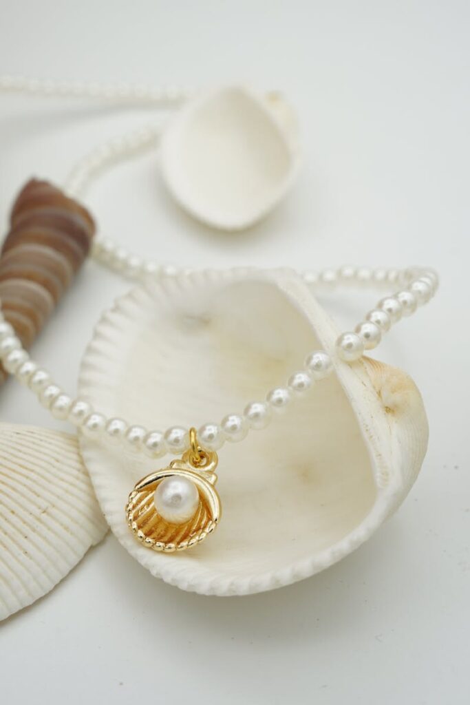 A close-up of a shimmering pearl chain with a gold seashell pendant, elegantly resting among seashells.