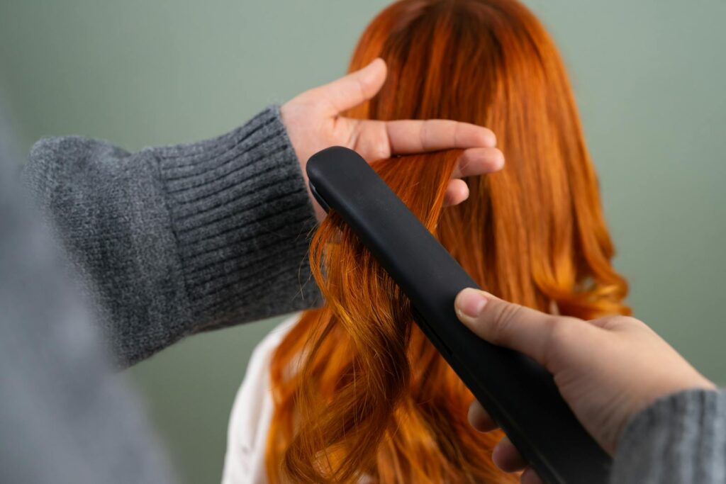 Hands styling long red hair using a flat iron indoors. Haircare and beauty concept.