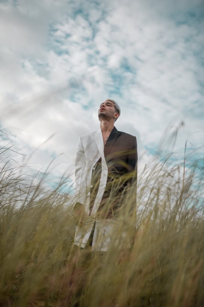 A man posing fashionably in a field with a cloudy sky backdrop, capturing a sense of freedom.