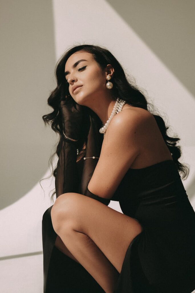 Woman in elegant black dress with pearl jewelry, posing artistically indoors.