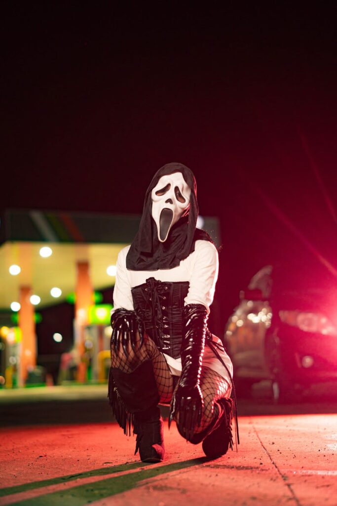 A person in a ghost mask kneels at a gas station at night, with vibrant lights enhancing the eerie atmosphere.