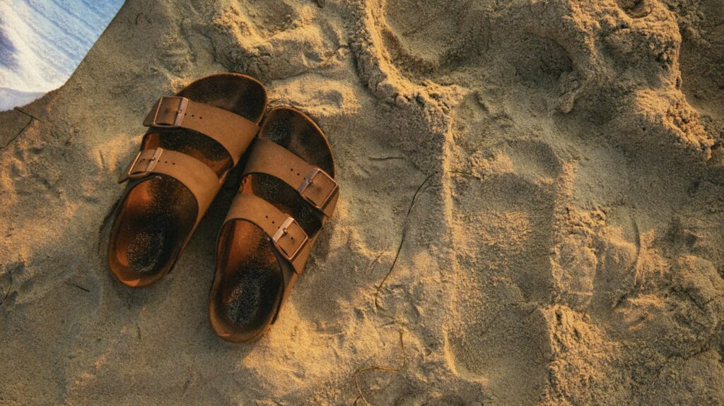 Top view of leather sandals on a sandy beach during sunrise, exuding a warm vacation vibe.