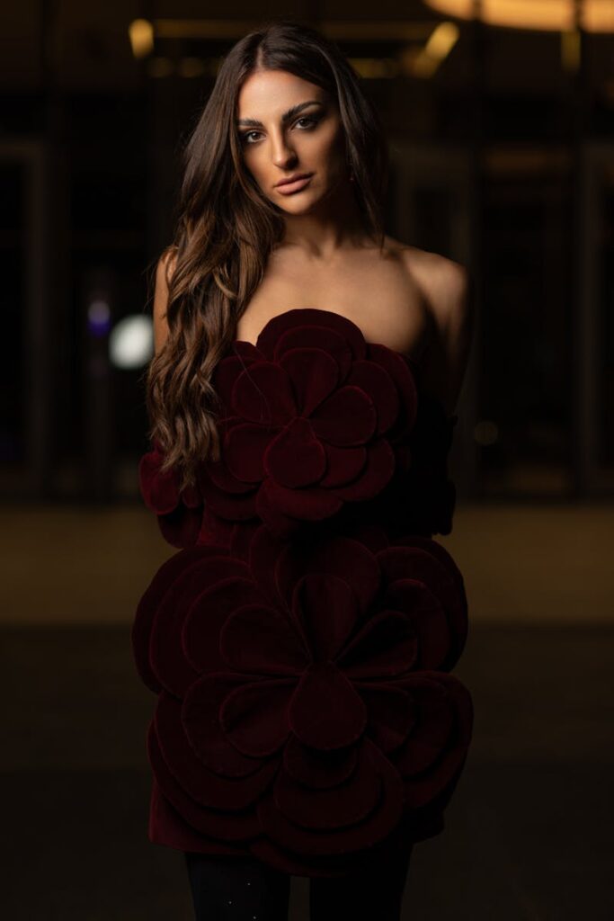 A beautiful young woman poses elegantly in a burgundy floral dress during nighttime.