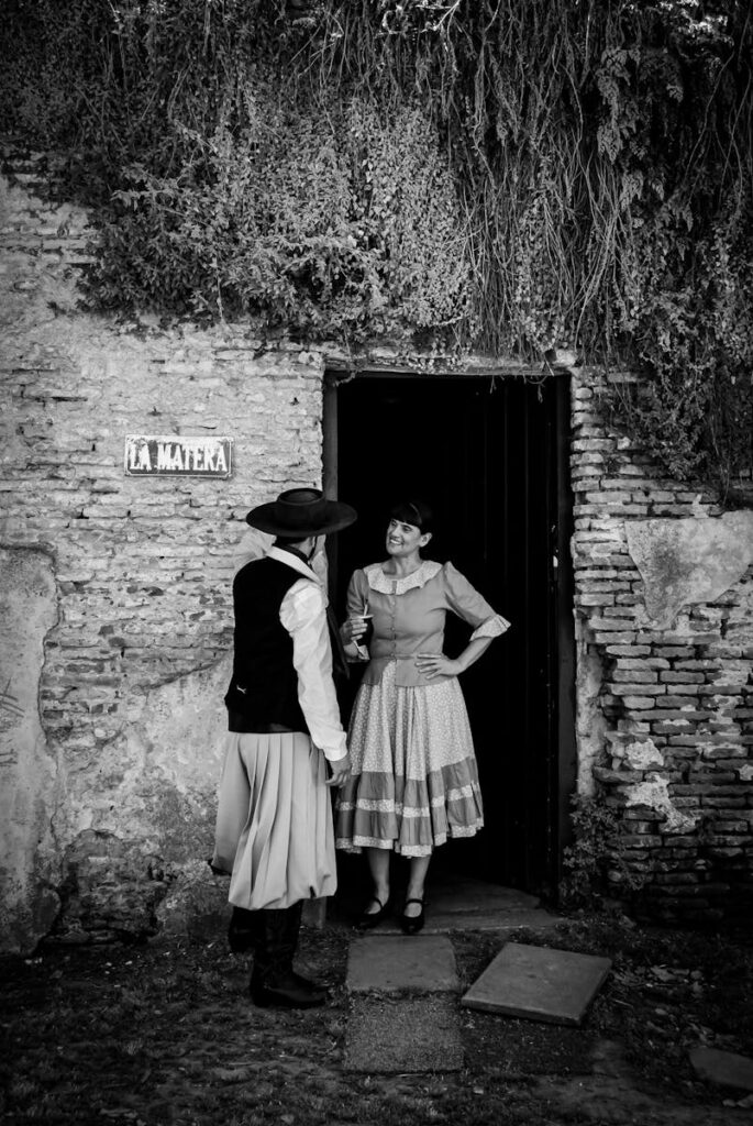 A man and woman in traditional attire converse outside a rustic stone building.