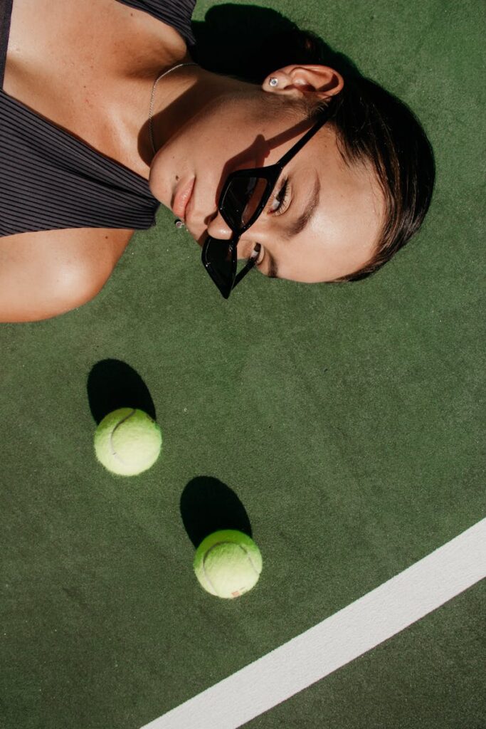 Woman in sunglasses lying on a tennis court with two tennis balls on a sunny day.