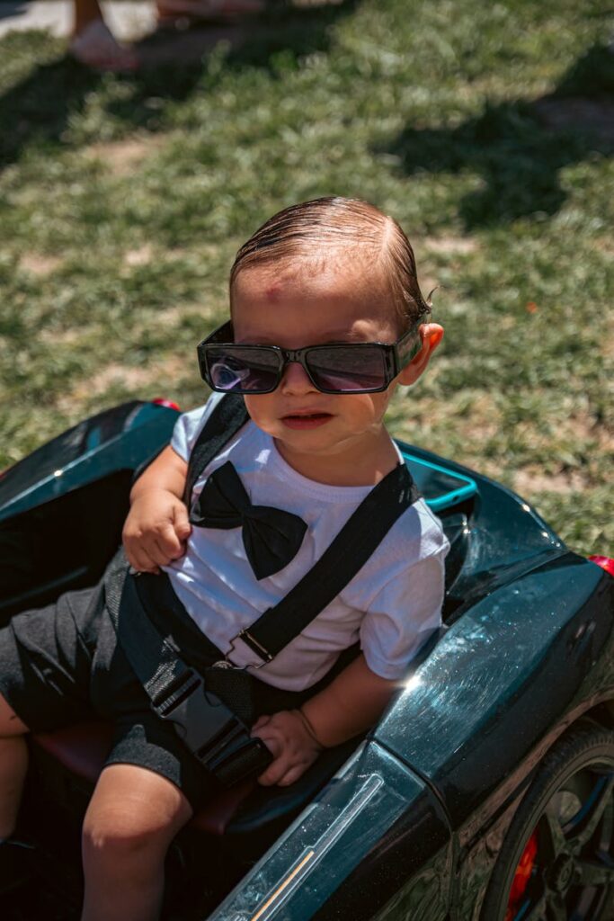 Adorable baby wearing sunglasses and formal attire sitting in a toy car outdoors.