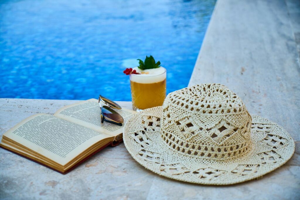 Enjoy a tranquil summer moment with a refreshing drink, hat, and book by the poolside.