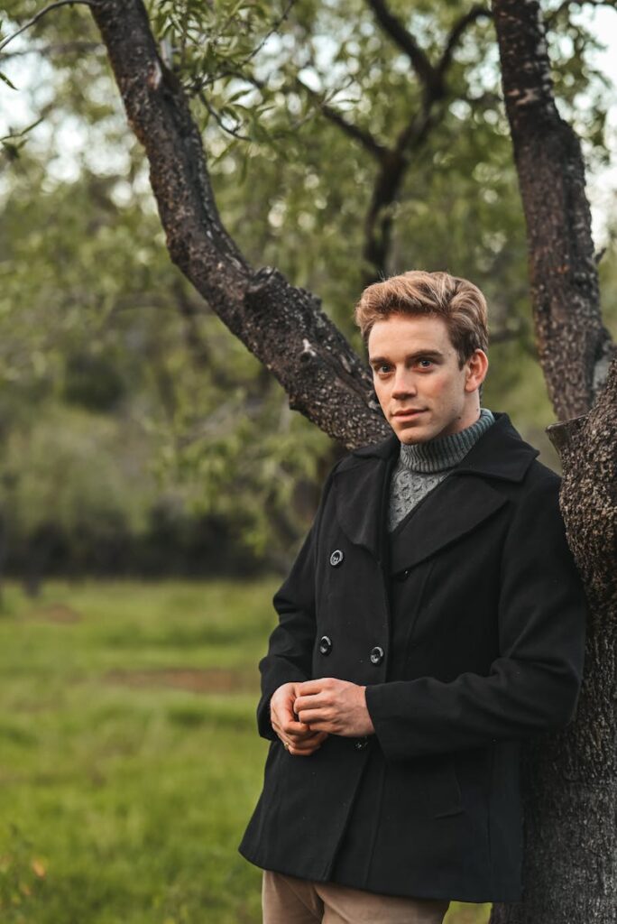 A young man leans against a tree outdoors in fall, wearing a black peacoat.