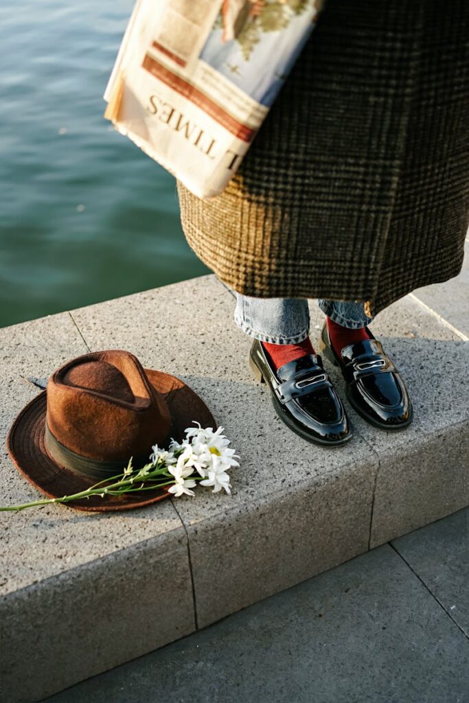 Elegant shoes near water, with a hat, newspaper, and flowers on a sunny day.