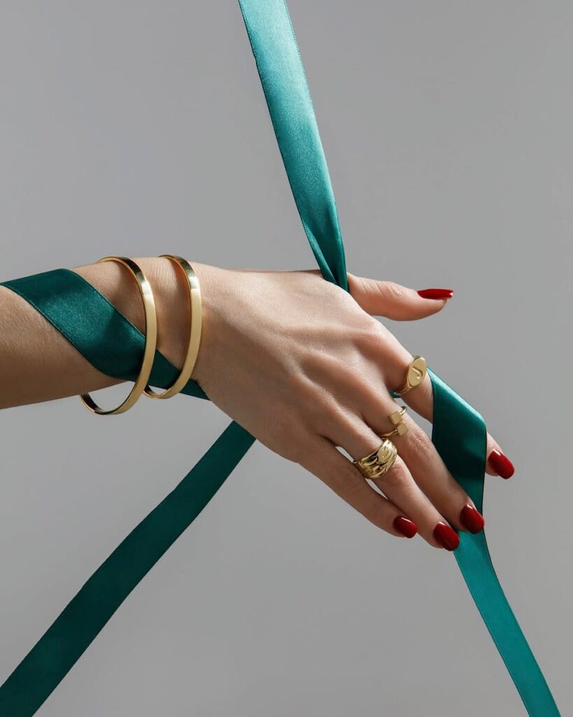 Close-up of elegant hands with gold jewelry and teal ribbon against a neutral background.