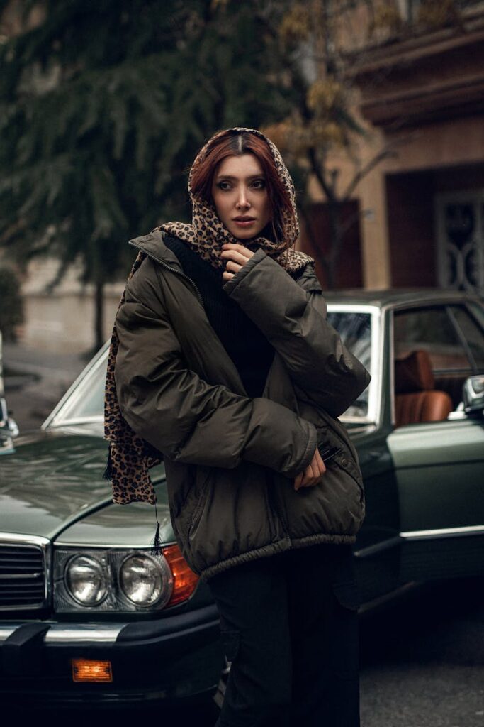 Fashionable woman in winter coat poses next to a vintage car in an urban setting.