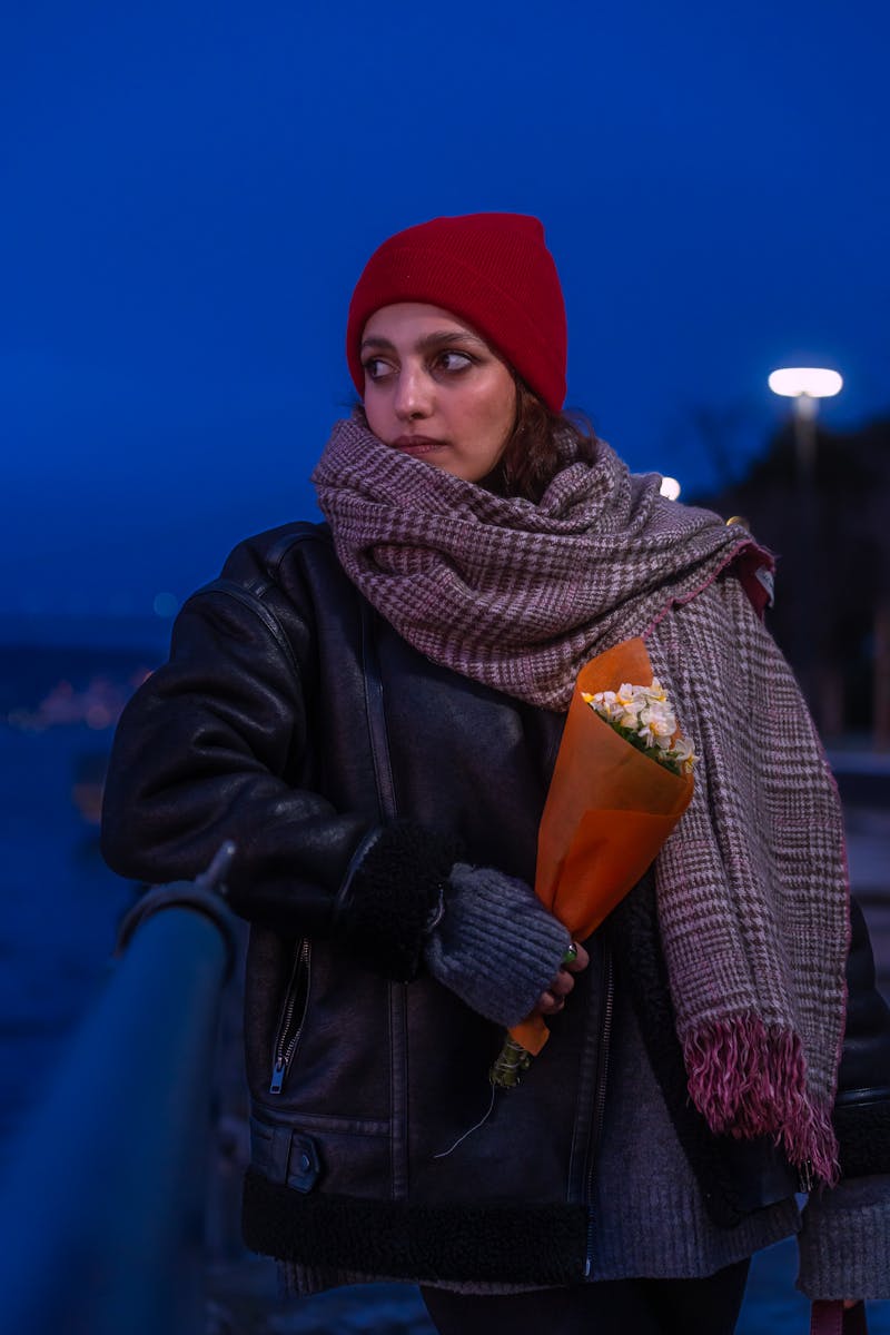 Woman in a red beanie and scarf holding flowers on a cold evening. Moody and vibrant.