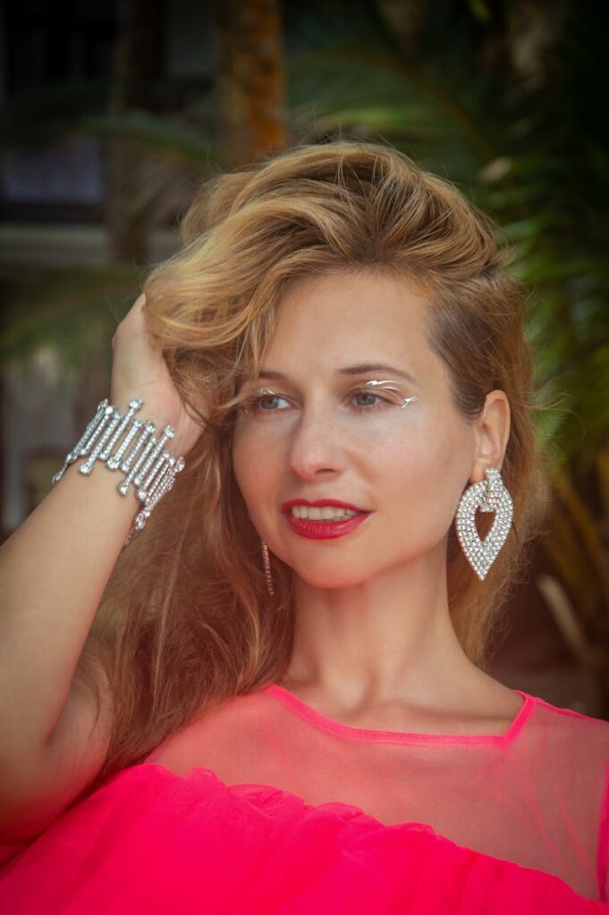 Elegant woman with diamond jewelry poses gracefully in a tropical setting wearing vibrant pink attire.