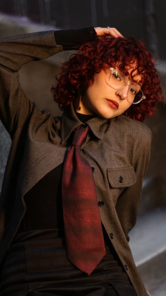 Young woman with curly hair and glasses in stylish attire strikes a pose outdoors, embodying modern fashion.