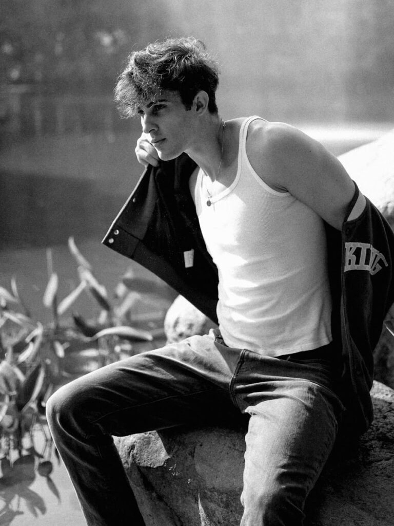 A young man in a white tank top and jeans sits pensively on a rock in a natural setting.