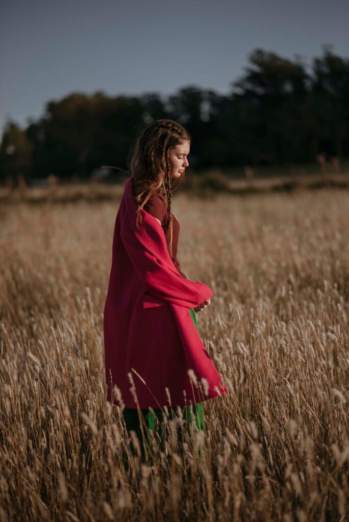 A contemplative woman in a red coat strolls through a serene autumn wheat field at sunset.