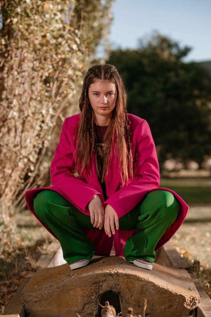 A woman with long hair in a pink coat and green pants poses outdoors.