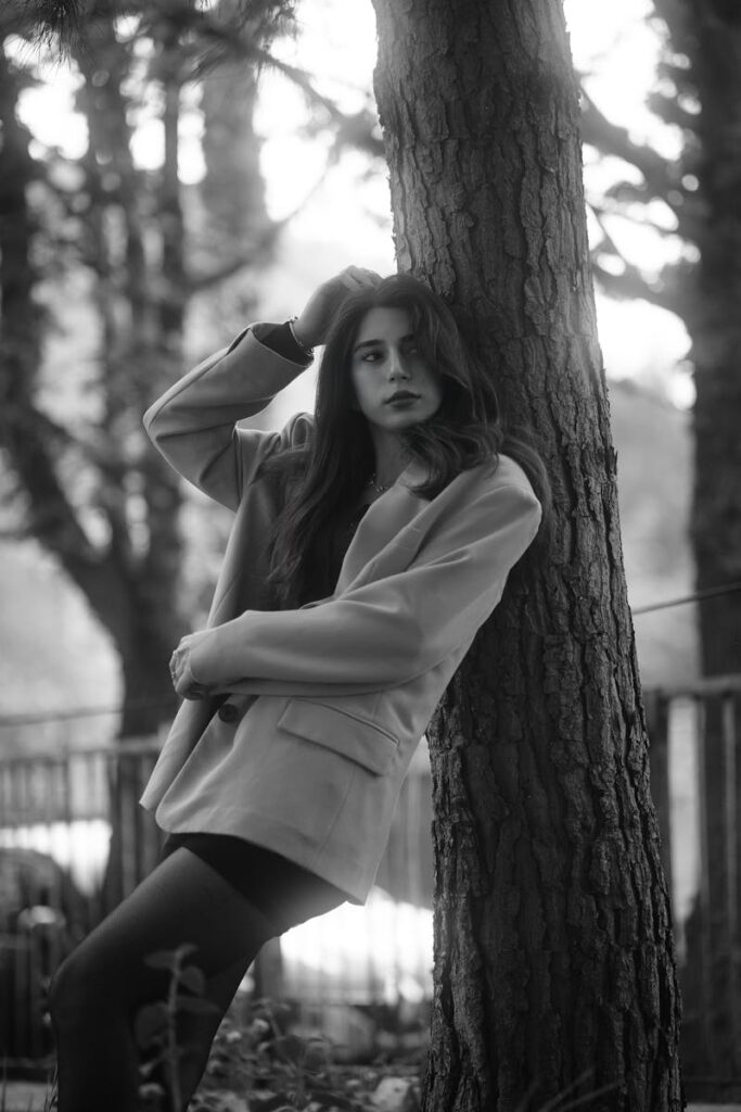 Black and white portrait of a woman leaning against a tree in a tranquil natural setting.