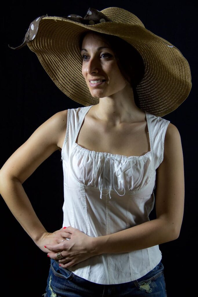 Portrait of a smiling woman in a wide-brimmed sun hat and white top against a dark background.
