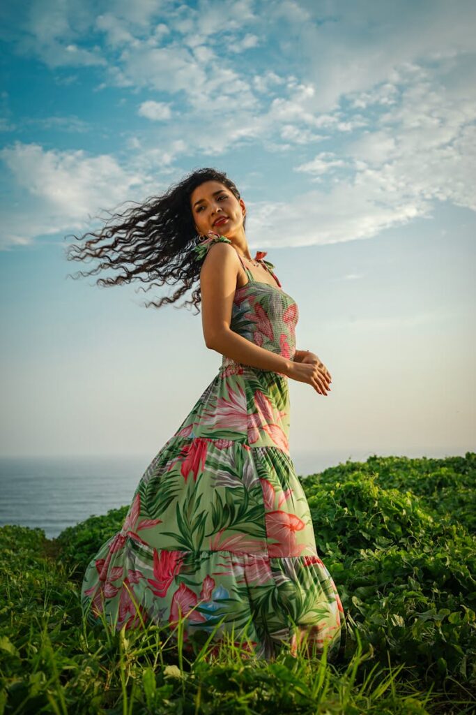 A woman in a floral dress enjoys a sunny day on a grassy coastal hill with a vibrant blue sky.