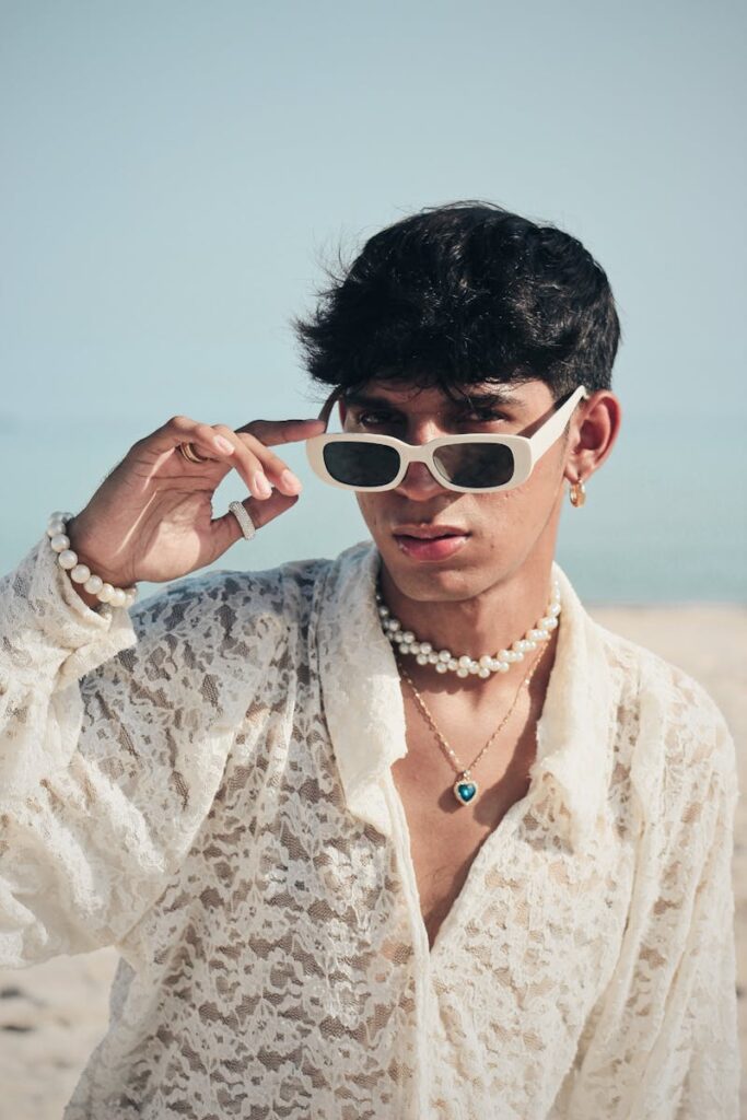 Stylish young man in lace shirt and sunglasses poses confidently on a sunny beach.