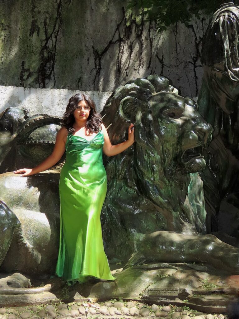 Elegant woman in a green dress poses by a majestic lion statue outdoors.